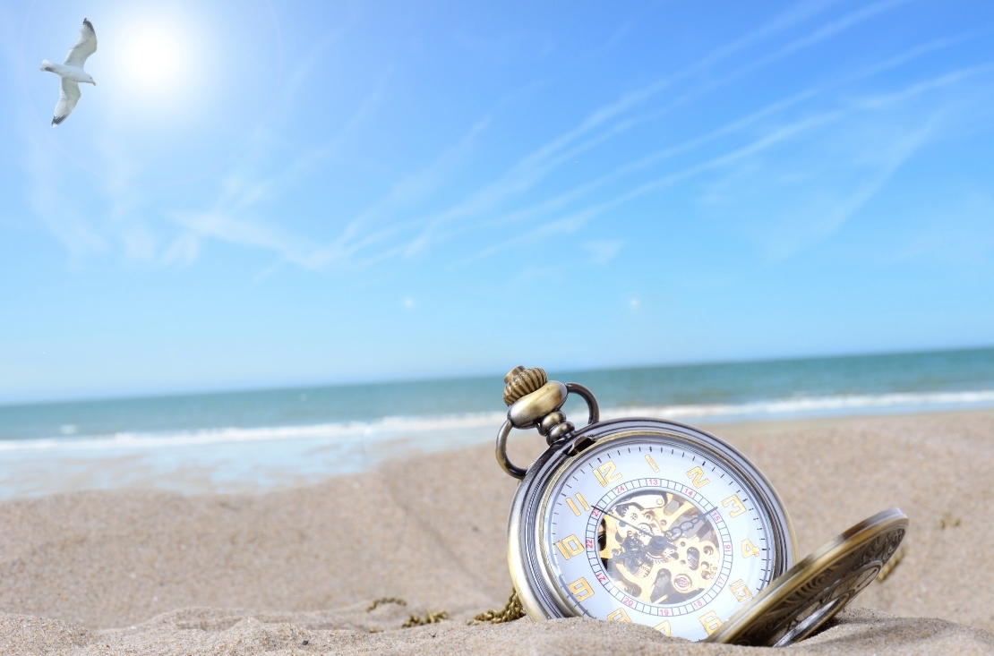 pocket watch in sand on a beach with water in the background to indicate a daylight savings discount on a beach vacation in Gulf Shores and Orange Beach