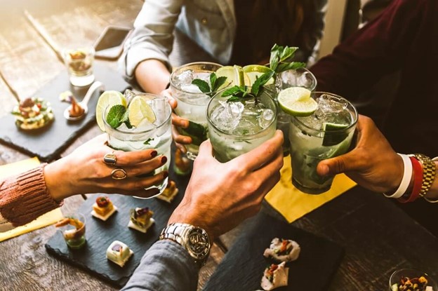 Group of four friends at a table raising a toast with iced cocktails garnished with lime and mint leaves.