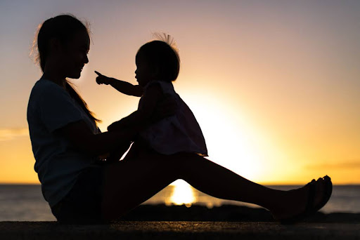 Silhouette of a mother sitting on a beach holding her young child up at sunset.