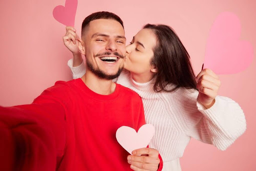 Woman kissing a man on the cheek while taking a selfie together against a pink background with heart decorations.