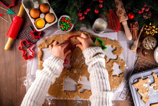 Overhead view of someone in a cozy sweater using star-shaped cookie cutters on dough, surrounded by baking ingredients, gingerbread men, and festive holiday decor.