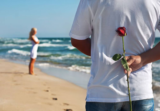 Man holding a red rose behind his back on a sandy beach while looking towards a woman in the distance near the ocean waves.